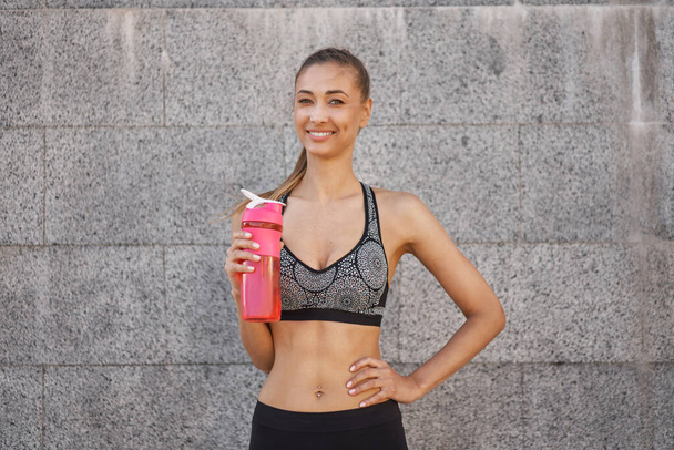 Tired Woman drink water red bottle after morning workout Young athletic female rear view standing leaning wall city street park background after jogging Healthy lifestyle concept Copy space - Foto, Imagen