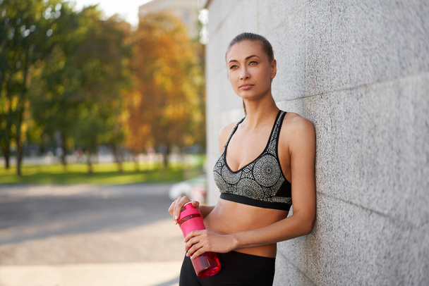 Tired Woman drink water red bottle after morning workout Young athletic female rear view standing leaning wall city street park background after jogging Healthy lifestyle concept Copy space - Фото, зображення