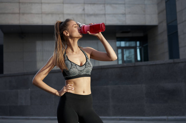 Tired Woman drink water red bottle after morning workout. Young athletic femalestanding city street background after jogging Healthy lifestyle concept  - Φωτογραφία, εικόνα