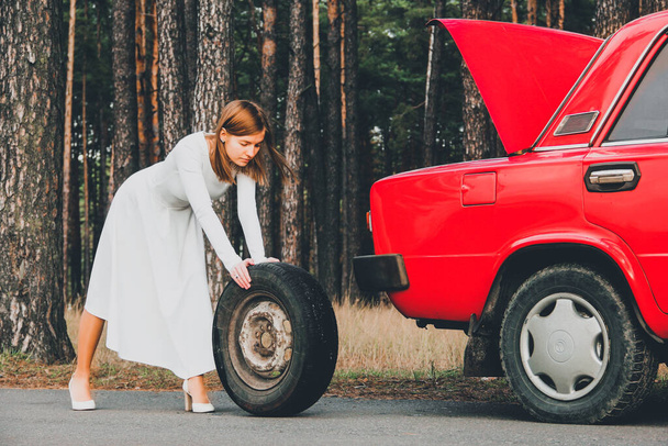 Chernigov, Ukraine - November 10, 2020: Girl in a wedding dress rolls the wheel. Vaz 2101. Red retro car in the forest - Foto, Imagem