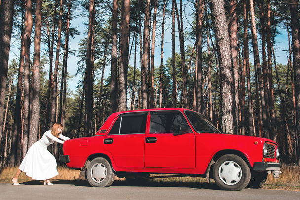 Chernigov, Ukraine - November 10, 2020: A girl in a wedding dress is pushing a car. Vaz 2101. Red retro car in the forest - Foto, immagini