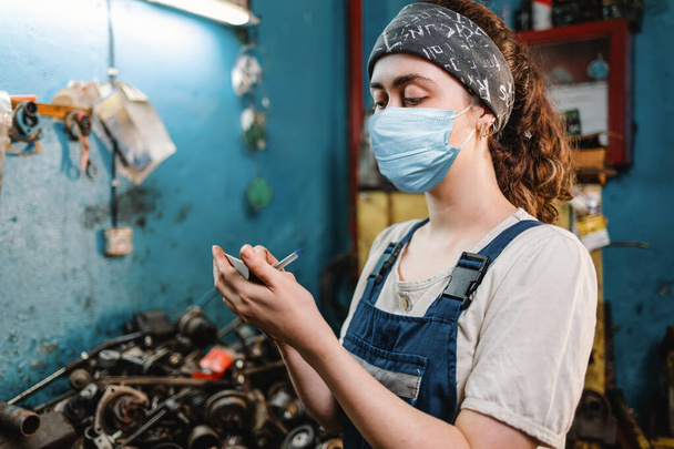 Gender equality. Portrait of a young woman in uniform, working in a workshop, which writes down orders on a piece of paper. In the background is a blue wall and a table with spare parts for the car. Close up. - Photo, Image