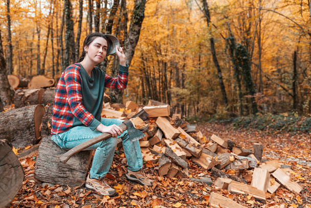 Voorbereiding van het verwarmingsseizoen. Een jonge mooie vrouw met een bijl in haar handen zit op een boomstam en doet haar pet af. Er is een stapel brandhout in de buurt.. - Foto, afbeelding