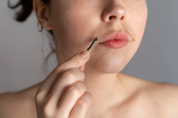 Eine junge Frau mit Schnurrbart versucht, die Haare über ihrer Lippe mit einer Pinzette zu entfernen. Das Konzept, unerwünschte Gesichtsbehaarung loszuwerden. Nahaufnahme - Foto, Bild