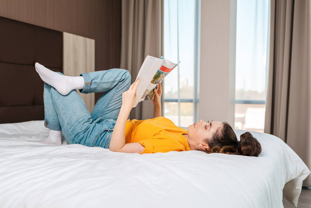 Lezen en onderwijs. Een jonge vrouw leest een tijdschrift liggend op het bed. Binnenland. Zijaanzicht. - Foto, afbeelding