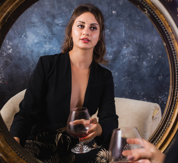 Gyönyörű fiatal hölgy fekete kabátban ül afrontján a vintage tükör üveg bor stúdió portré. - Fotó, kép