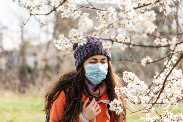 Eine Frau in medizinischer Maske steht auf der Straße neben einem blühenden Baum und versucht, die Blumen zu riechen. Konzept der saisonalen Pollenallergie und Virusinfektionen. - Foto, Bild