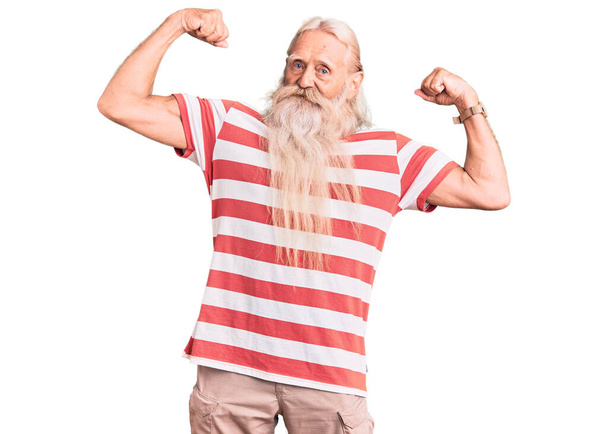 Vieil homme âgé aux cheveux gris et à la barbe longue portant un t-shirt rayé montrant les muscles des bras souriant fier. concept de fitness.  - Photo, image
