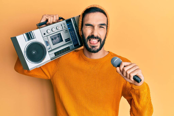 Young hispanic man with beard holding boombox listening to music singing with microphone angry and mad screaming frustrated and furious, shouting with anger. rage and aggressive concept.  - Фото, зображення