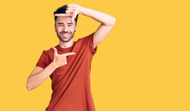 Joven hombre hispano vistiendo ropa casual sonriendo haciendo marco con manos y dedos con cara feliz. concepto de creatividad y fotografía.  - Foto, imagen