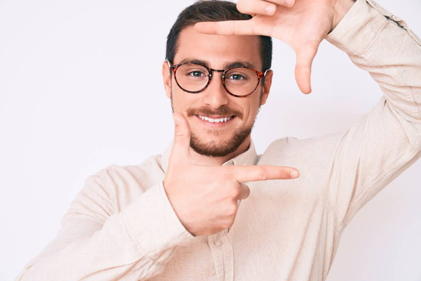 Jonge knappe man met casual kleding en een glimlachende bril die een frame maakt met handen en vingers met een vrolijk gezicht. creativiteit en fotografie.  - Foto, afbeelding