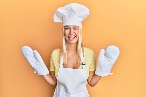 Young blonde girl wearing baker uniform and cook hat winking looking at the camera with sexy expression, cheerful and happy face.   - Photo, Image