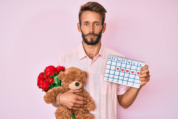 Handsome caucasian man with beard holding bear and bouquet of flowers for anniversary relaxed with serious expression on face. simple and natural looking at the camera.  - Photo, Image