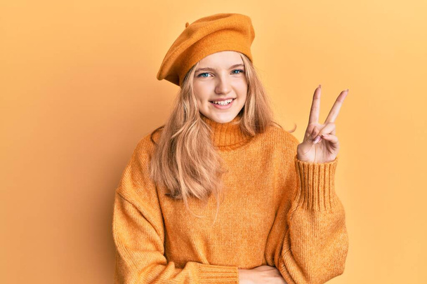 Beautiful young caucasian girl wearing french look with beret smiling with happy face winking at the camera doing victory sign. number two.  - Foto, immagini