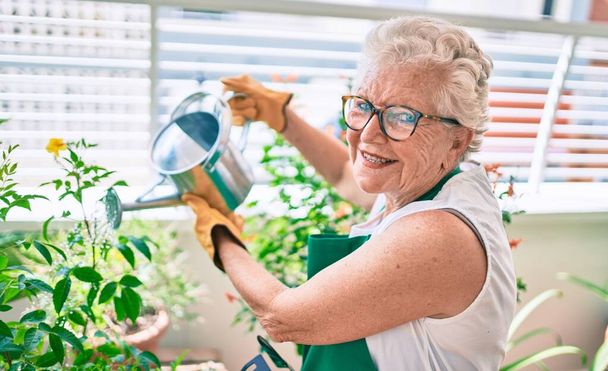 Senior woman with grey hair wearing gloves and gardener apron gardening the plants at home - Foto, imagen