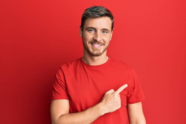 Hombre caucásico guapo vistiendo camiseta roja casual alegre con una sonrisa en la cara señalando con la mano y el dedo hacia el lado con expresión feliz y natural  - Foto, Imagen