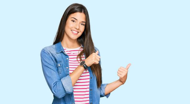 Young brunette woman wearing casual clothes pointing to the back behind with hand and thumbs up, smiling confident  - 写真・画像