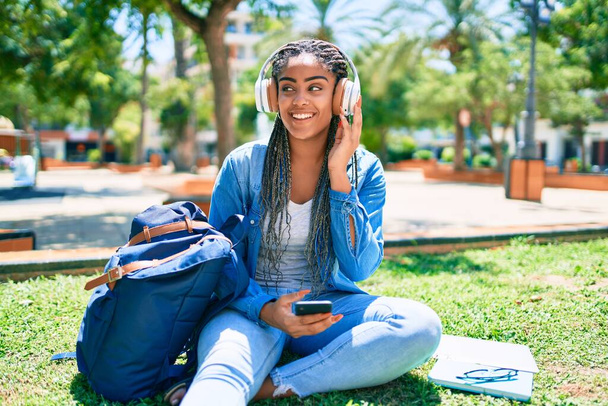 若いアフリカ系アメリカ人の学生の女性が笑顔で大学のキャンパスで草の上に座って音楽を聞いて幸せ - 写真・画像