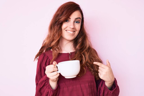 Mladá krásná žena drží kávu s úsměvem šťastný ukazující rukou a prstem  - Fotografie, Obrázek
