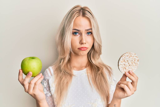 Piękna biała blondynka trzyma nachos i zdrowe zielone jabłko przygnębiony i martwić się o stres, płacz zły i przerażony. smutna ekspresja.  - Zdjęcie, obraz