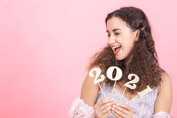 Schöne elegante junge brünette Mädchen mit lockigem Haar und nackten Schultern hält eine hölzerne Zahl 2021 in ihren Händen auf einem rosa Hintergrund, Neujahrsurlaub Konzept - Foto, Bild