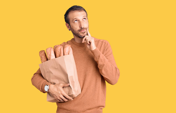 Молодой красивый мужчина держит бумажный пакет с хлебом серьезное лицо думая о вопросе с рукой на подбородке, вдумчивый о запутанной идее  - Фото, изображение