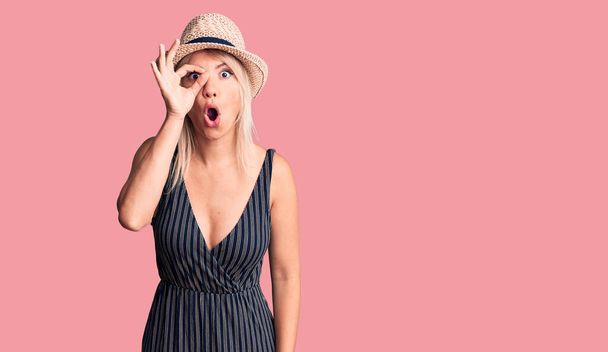 Jonge mooie blonde vrouw dragen zomer hoed en jurk doen ok gebaar geschokt met verrast gezicht, oog kijken door vingers. ongelovige uitdrukking.  - Foto, afbeelding
