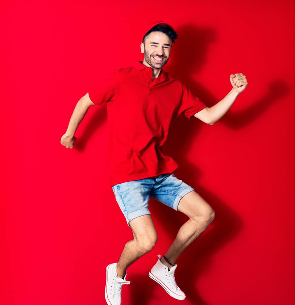 Young handsome deliveryman wearing cap smiling happy. Jumping with smile on face over isolated red background - Zdjęcie, obraz