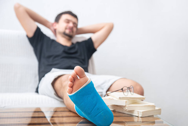 Mężczyzna ze złamaną nogą w niebieskiej szynie do leczenia urazów po skręceniu kostki czytanie książek w domu rehabilitacji. - Zdjęcie, obraz