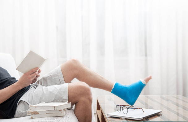 Um homem com uma perna quebrada em um elenco lê livros contra um fundo leve do interior da sala. - Foto, Imagem
