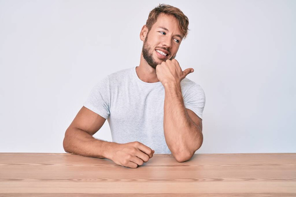 Jonge blanke man draagt casual kleding zittend op de tafel glimlachend met een vrolijk gezicht kijkend en wijzend naar de zijkant met duim omhoog.  - Foto, afbeelding