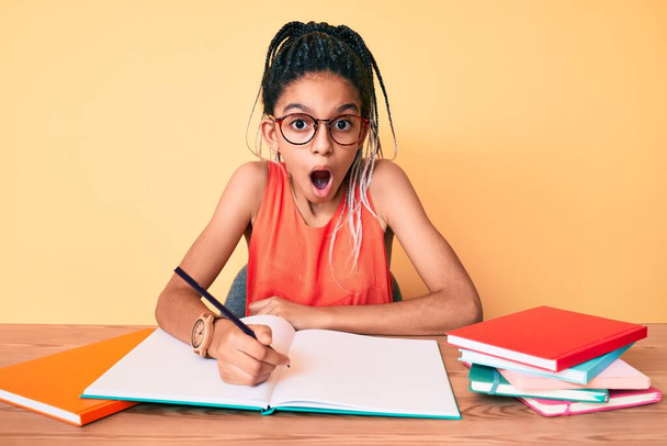 Jovem menina afro-americana criança com tranças estudando para o exame escolar assustado e surpreso com a boca aberta para surpresa, rosto descrença  - Foto, Imagem