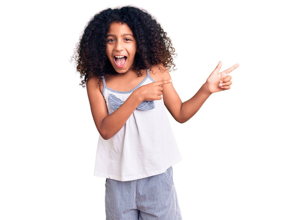 African american child with curly hair wearing casual clothes smiling and looking at the camera pointing with two hands and fingers to the side.  - 写真・画像