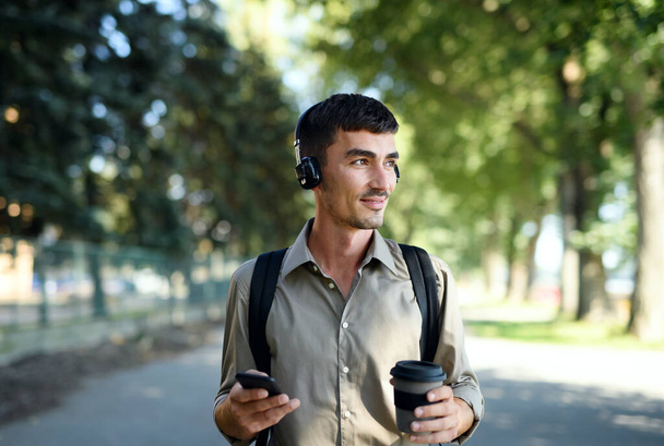 Jonge man met koptelefoon en koffie in herbruikbare beker wandelen buiten in de stad. - Foto, afbeelding