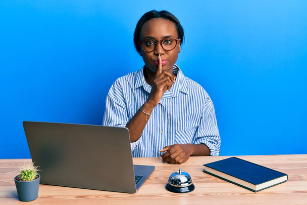 Młoda Afrykanka pracująca w recepcji za pomocą laptopa prosząc o ciszę z palcem na ustach. cisza i tajne pojęcie.  - Zdjęcie, obraz