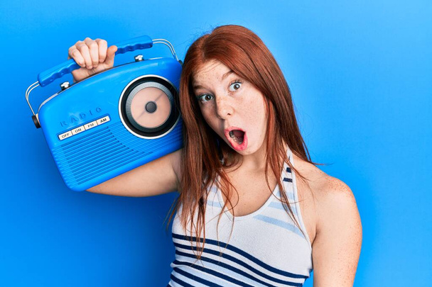 junges Mädchen mit rotem Kopf hält Vintage Radio verängstigt und erstaunt mit offenem Mund für Überraschung, ungläubiges Gesicht  - Foto, Bild