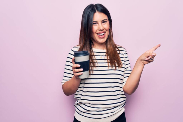 Mladá krásná brunetka žena pití šálek kávy přes izolované růžové pozadí s úsměvem šťastný ukazující rukou a prstem na stranu - Fotografie, Obrázek