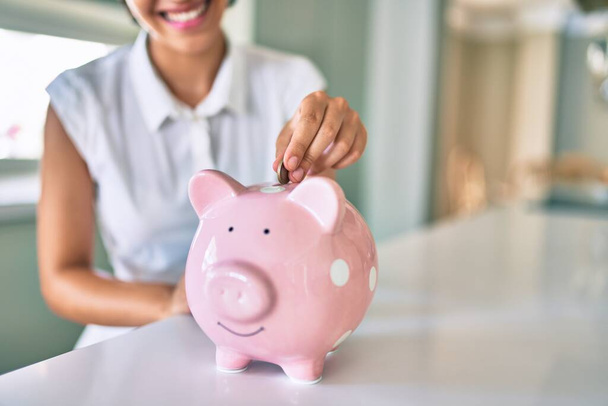 Junge brünette Frau lächelt glücklich und legt Ersparnisse in Sparschwein - Foto, Bild