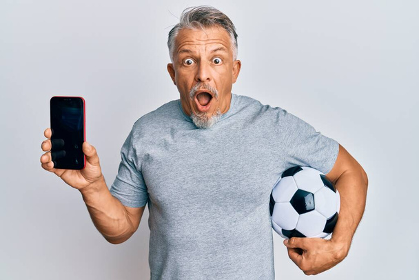 Μέση ηλικία γκριζομάλλης άνδρας κρατώντας μπάλα ποδοσφαίρου δείχνει smartphone φοβισμένος και σοκαρισμένος με έκπληξη και κατάπληκτος έκφραση, φόβο και ενθουσιασμένος πρόσωπο.  - Φωτογραφία, εικόνα