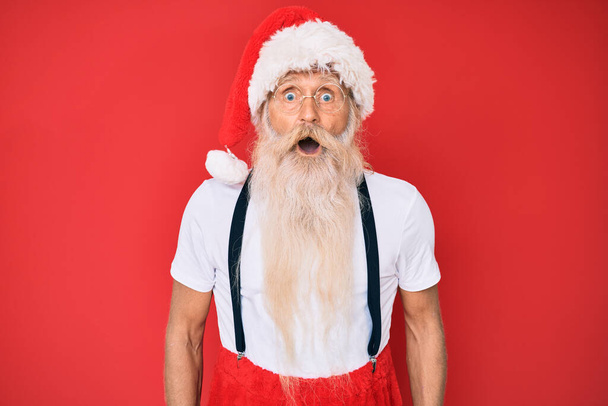 Staruszek z siwymi włosami i długą brodą w białej koszulce i kostiumie Świętego Mikołaja przestraszony i zszokowany zaskakującą ekspresją, strachem i podnieconą twarzą.  - Zdjęcie, obraz