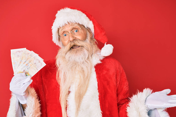 Staruszek z siwymi włosami i długą brodą w kostiumie Świętego Mikołaja, trzymający funty świętujące osiągnięcie ze szczęśliwym uśmiechem i wyrazem zwycięzcy z podniesioną ręką.  - Zdjęcie, obraz