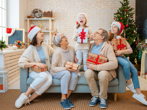 Merry Christmas and Happy Holidays! Grandma, grandpa, mum and children exchanging gifts. Parents and daughters having fun near tree indoors. Loving family with presents in room. - Photo, image