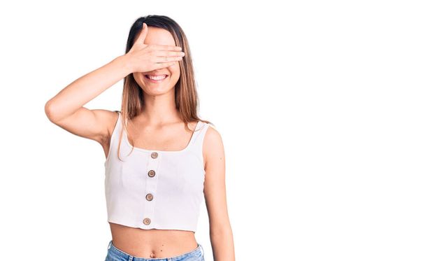 Young beautiful girl wearing casual sleeveless t shirt smiling and laughing with hand on face covering eyes for surprise. blind concept.  - Photo, Image