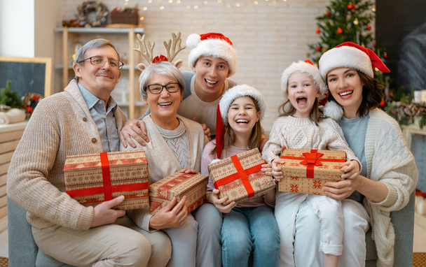 Merry Christmas and Happy Holidays! Grandma, grandpa, mum, dad and children exchanging gifts. Parents and daughters having fun near tree indoors. Loving family with presents in room. - Фото, изображение