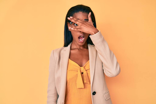 若いアフリカ系アメリカ人の女性は、顔や目を手で覆い、恥ずかしそうな表情で指を通して見てショックを受けてビジネス服を見ている.  - 写真・画像