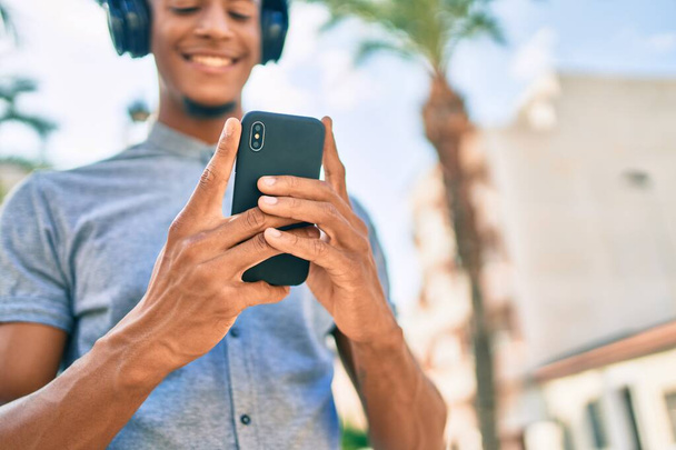 Νεαρός Αφροαμερικάνος χαμογελώντας χαρούμενος χρησιμοποιώντας smartphone και ακουστικά στην πόλη. - Φωτογραφία, εικόνα