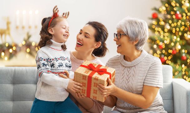 Merry Christmas and Happy Holidays! Cheerful kid presenting gifts to mom and granny. Parents and little child having fun near tree indoors. Loving family with presents in room. - Valokuva, kuva
