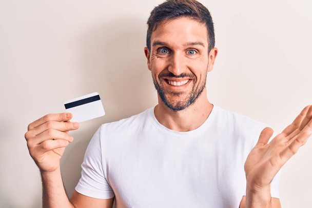 Young handsome customer man holding credit card over isolated white background celebrating achievement with happy smile and winner expression with raised hand - Photo, Image