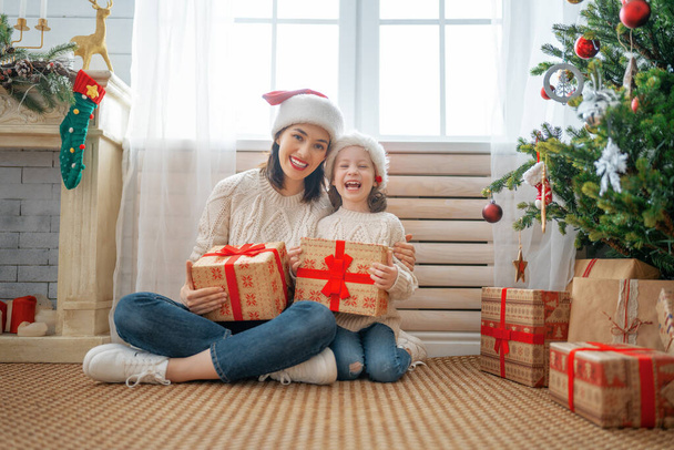 Merry Christmas and Happy Holidays! Cheerful mom and her cute daughter girl exchanging gifts. Parent and little child having fun near tree indoors. Loving family with presents in room.    - Photo, Image