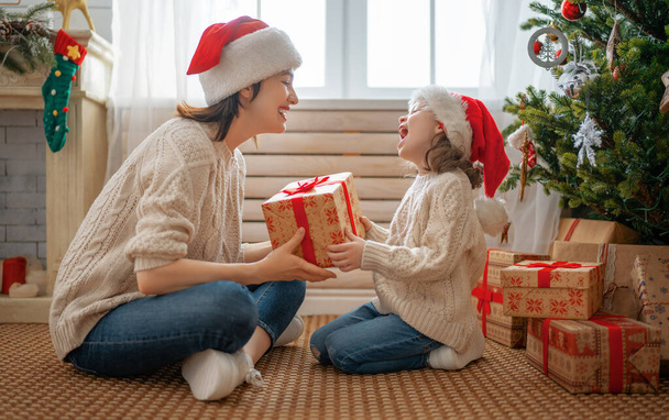 Merry Christmas and Happy Holidays! Cheerful mom and her cute daughter girl exchanging gifts. Parent and little child having fun near tree indoors. Loving family with presents in room.    - Foto, Imagem